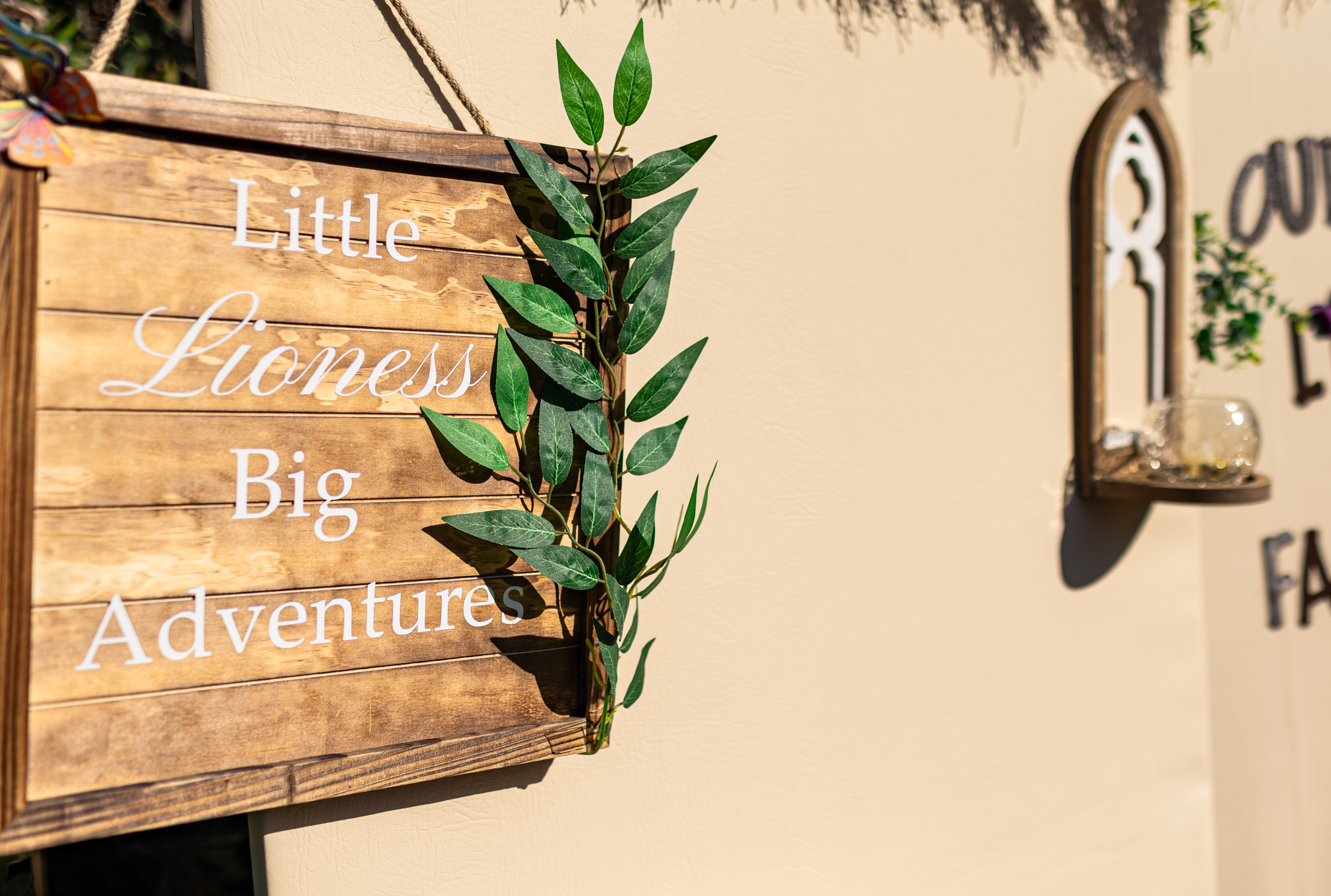Rustic wood-plank sign reading Little Lioness Big Adventures, dressed with trailing eucalyptus