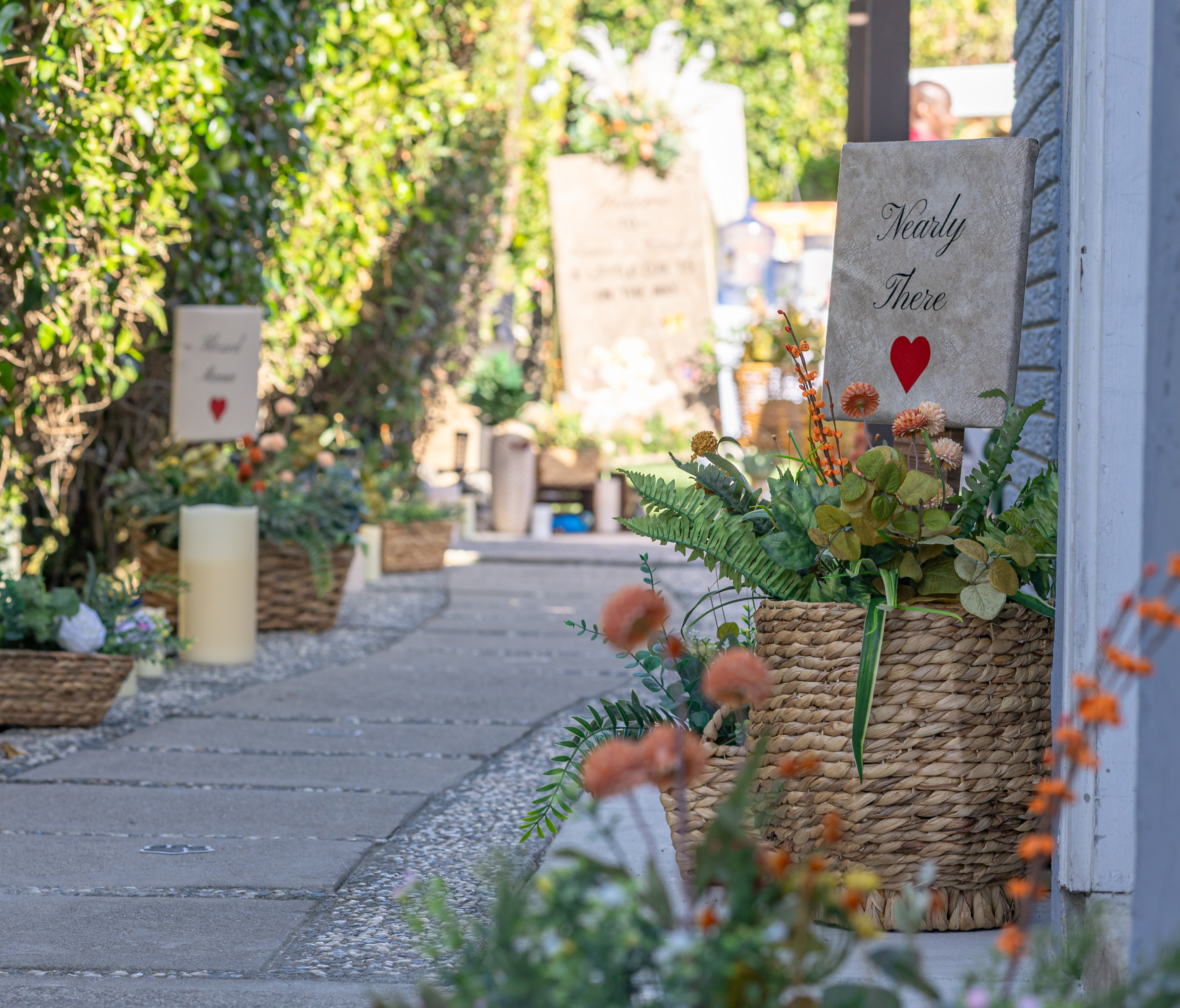 Flower-lined entrance pathway with wicker planters, ferns, and hand-lettered directional signs